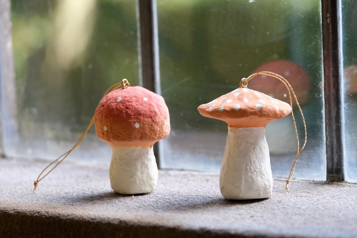 Two small mushroom-shaped ornaments with gold hanging strings, placed on a windowsill in front of a blurred window.