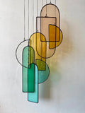 A stained glass sun catcher with multiple hanging glass pieces in various shapes and colors, including amber and teal, with a black patina finish and linen cord against a white background.