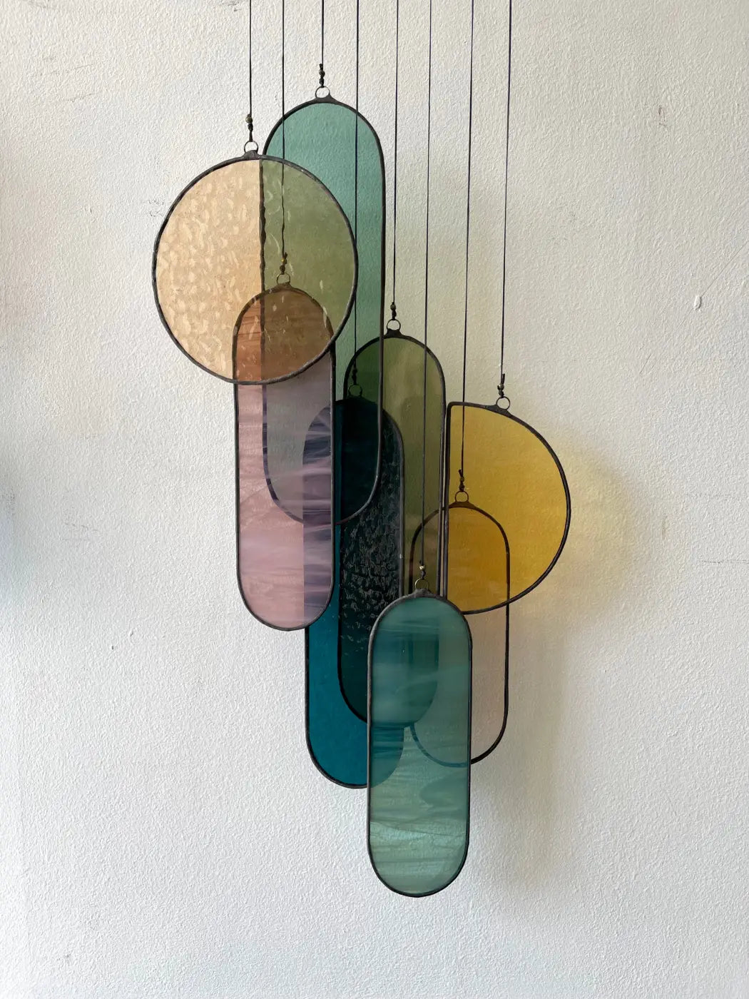 A wall-mounted stained glass sun catcher composed of multiple oval and circular pieces in various shades of blue, green, and amber, held together by a brass bar.