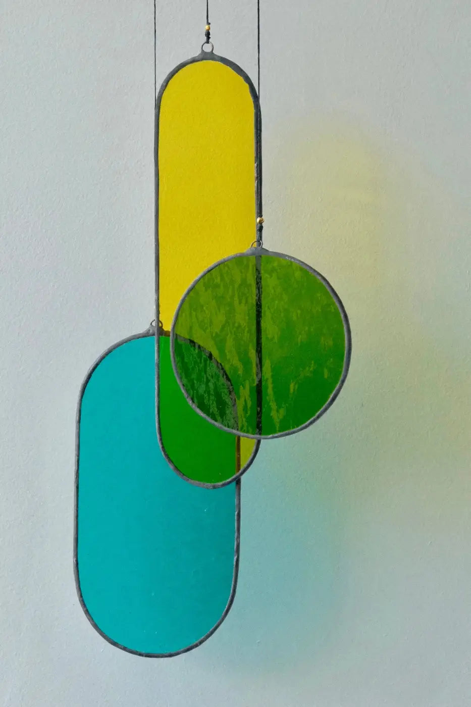 A hanging stained glass decoration featuring geometric shapes in green, yellow, and blue colors with a black patina finish, suspended by a brass hoop.