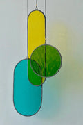 A hanging stained glass decoration featuring geometric shapes in green, yellow, and blue colors with a black patina finish, suspended by a brass hoop.