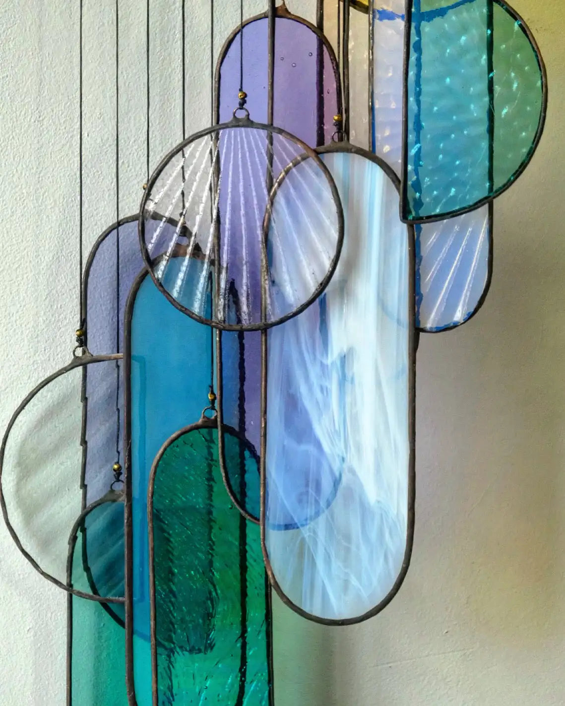 A colorful stained glass hanging art piece featuring oval and circular panels in shades of blue, green, purple, and clear glass with metallic frames.