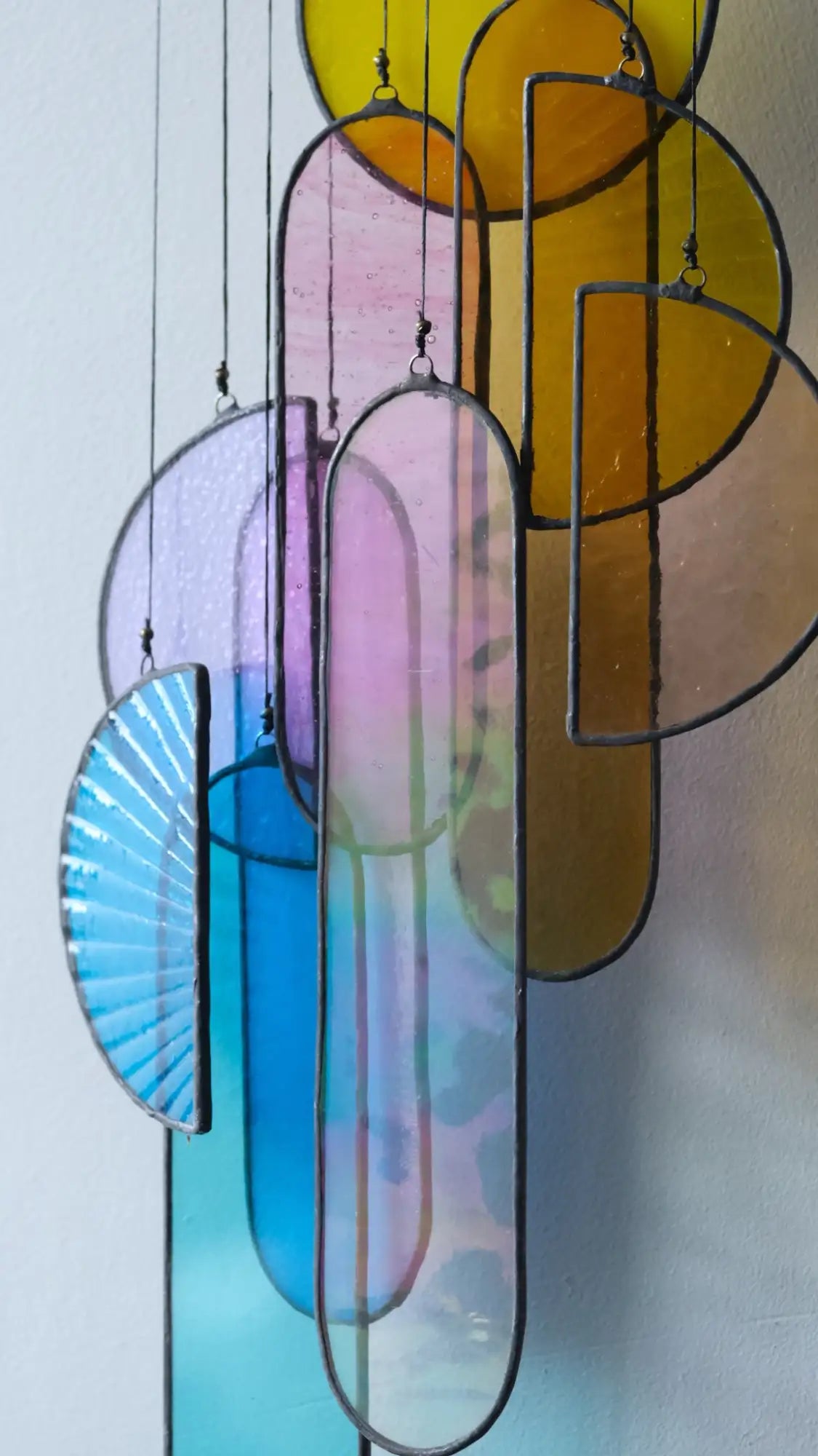 A colorful stained glass hanging art piece featuring translucent panels in shades of blue, purple, pink, and yellow with black metal framing.