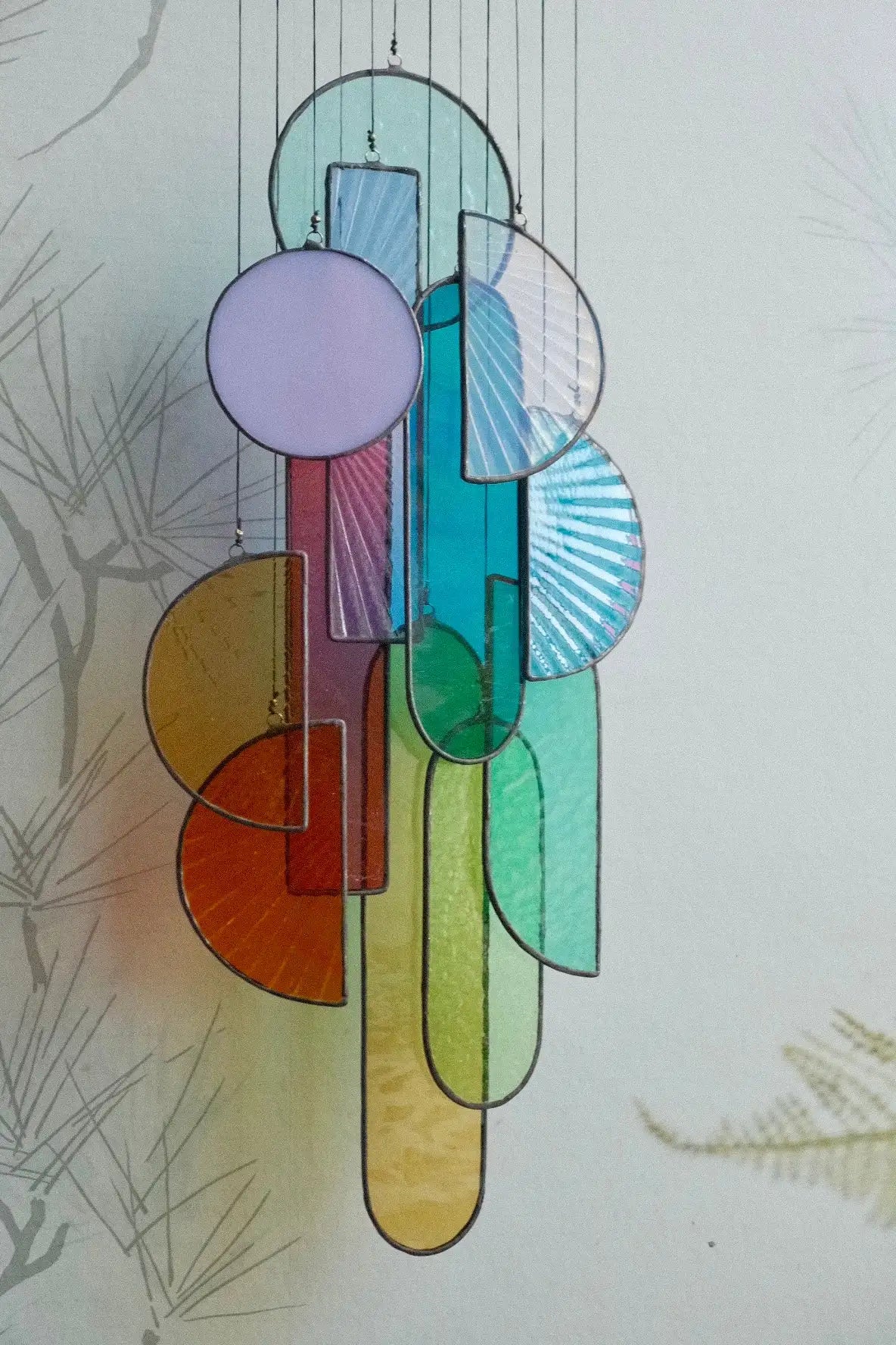 A colorful stained glass hanging mobile featuring geometric shapes in hues of lavender, teal, orange, and green.