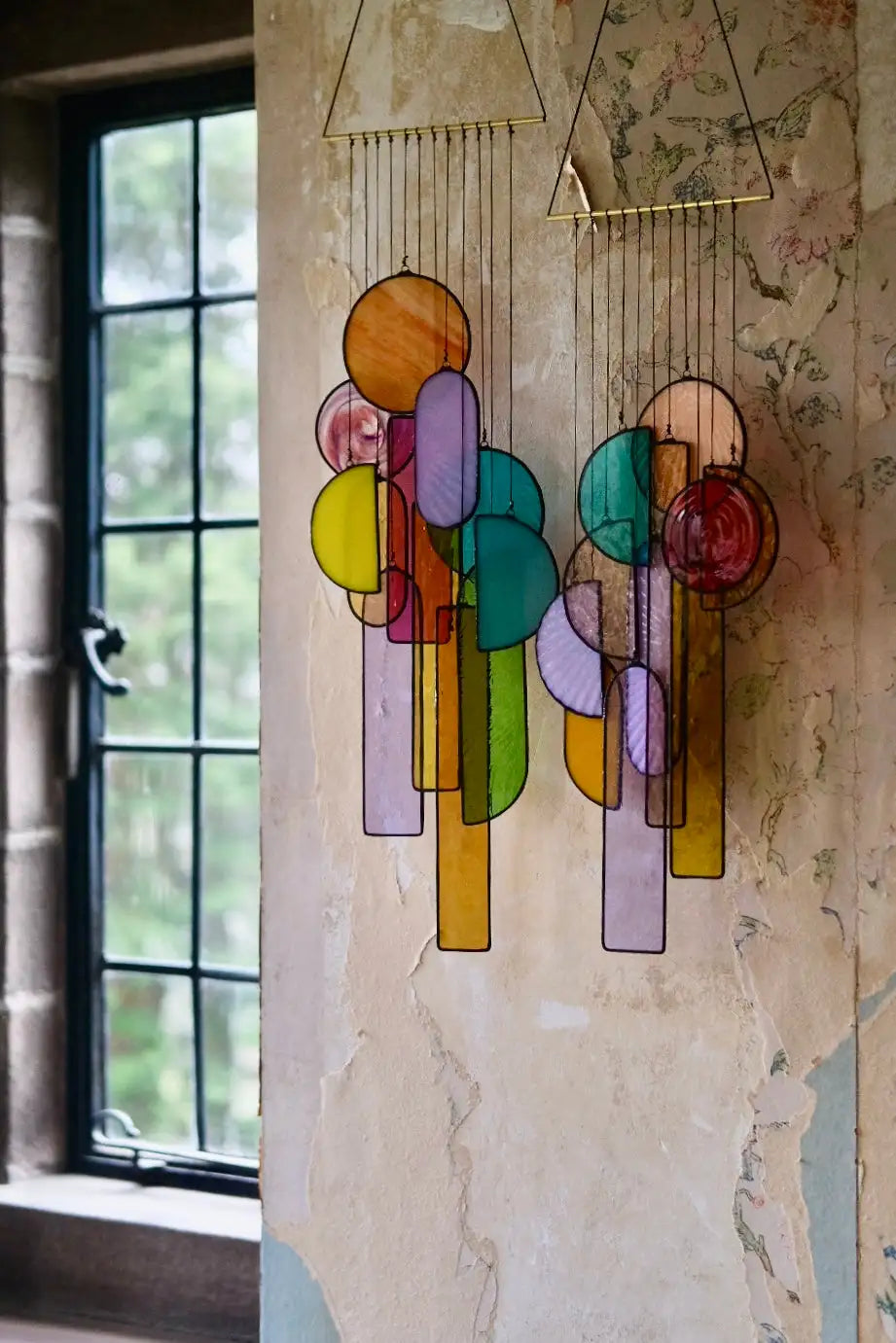 A colorful stained glass hanging mobile with geometric shapes in vibrant hues of yellow, green, purple, and orange suspended from gold-toned metal frames.