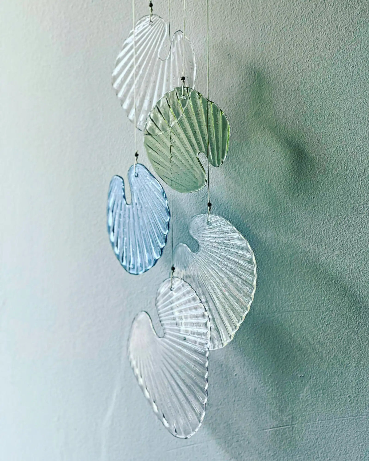 A delicate glass shell wind chime featuring translucent pieces in clear, soft green, and light blue hues with a subtle ribbed texture.