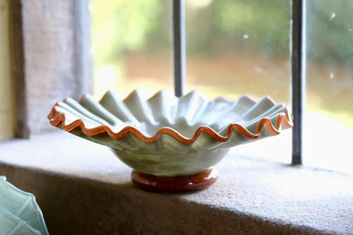 A glass bowl with a fluted, scalloped rim and a small round pedestal base, shown on a windowsill with a blurred outdoor background. The bowl has a pale sage green glaze with a contrasting terracotta brown rim and base.