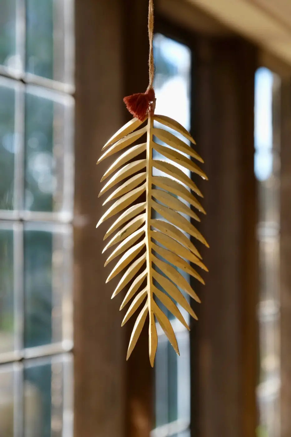 Handmade palm leaf decoration featuring a delicate golden-yellow wooden palm frond with a red tassel hanging from rustic twine