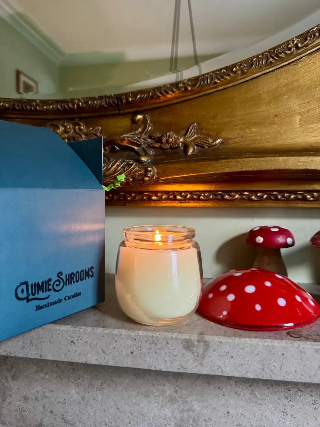 A creamy white scented candle in a clear glass jar with a lit wick next to red polka-dotted mushroom decor