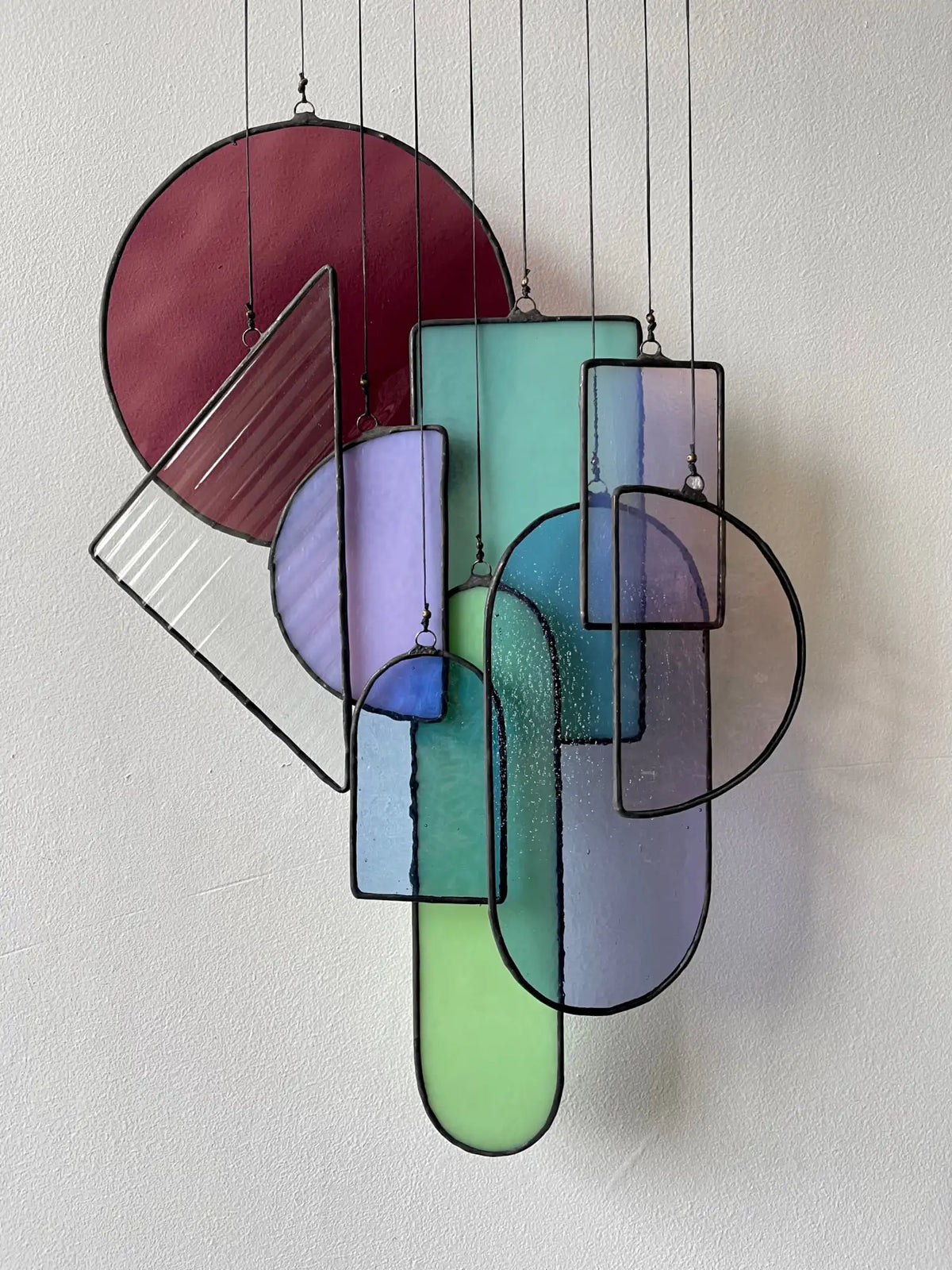 A modern stained glass hanging art piece featuring geometric shapes in vibrant hues of maroon, teal, lavender, and green with black leaded edges.