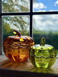 Two decorative glass pumpkins, one amber-orange and one light green, sitting on a windowsill with a window and outdoor scenery in the background.