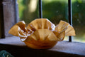 A handcrafted amber glass bowl with a ruffled, organic edge, resting on a dark windowsill with a blurred outdoor background.