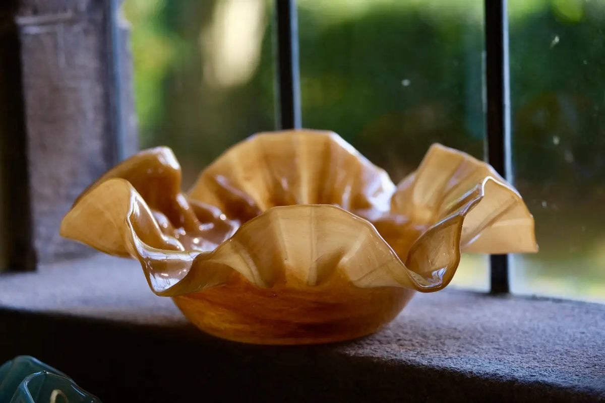 A handcrafted amber glass bowl with a ruffled, organic edge, resting on a dark windowsill with a blurred outdoor background.