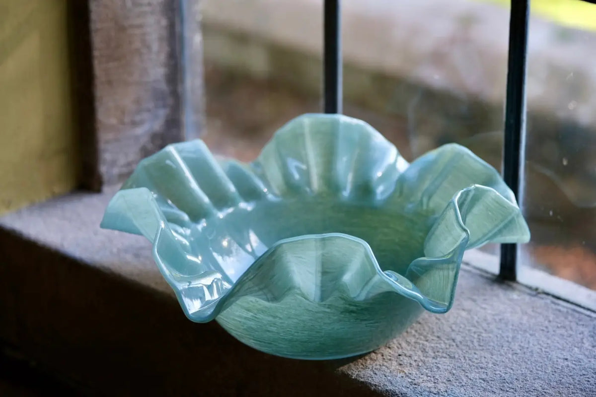 A light aqua green ruffled glass bowl resting on a stone windowsill with a window and bars in the background.