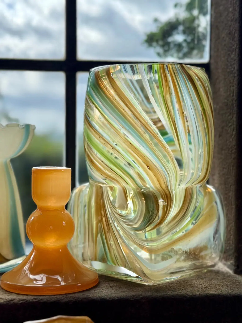 A hand-blown glass vase with swirling green, yellow, and white ribbons stands on a windowsill next to a small amber glass candle holder. A window with a cloudy sky is visible in the background.