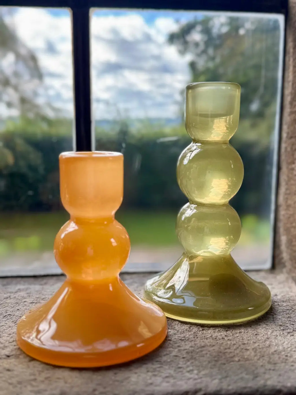 Two glass candlesticks with a bulbous tiered design, one in translucent amber-orange and the other in translucent pale yellow-green, displayed on a stone windowsill with a blurred outdoor garden background.