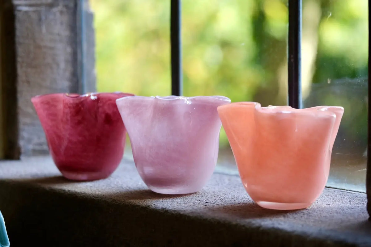 Three small flower-shaped glass candle holders in pink, red, and peach, lined up on a windowsill with a blurred green garden background.
