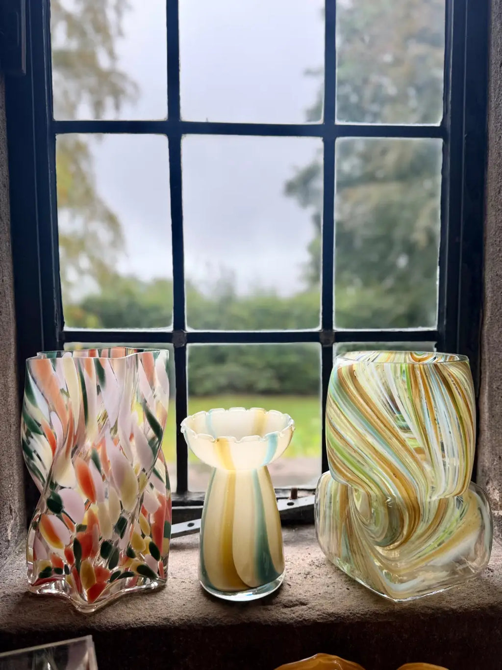 Three colorful hand-blown glass square swirl vases with swirling and floral patterns on a windowsill