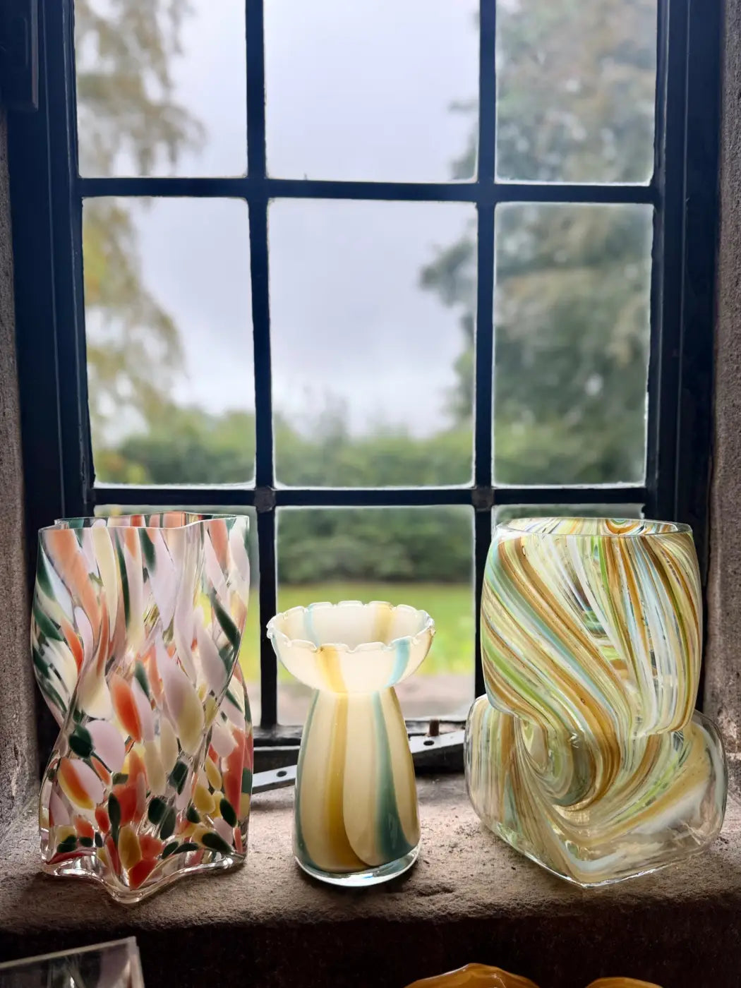 Three colorful hand-blown glass square swirl vases with swirling and floral patterns on a windowsill
