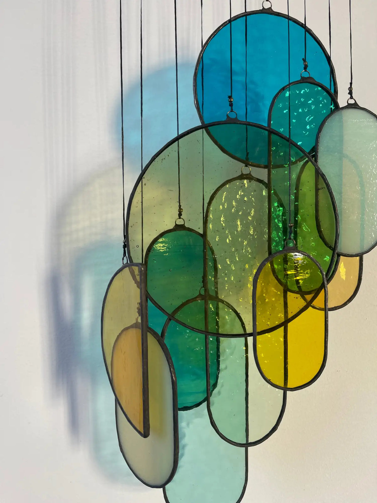 A hanging stained glass art piece featuring circular and oval shapes in vibrant shades of blue, green, yellow, and teal, framed with dark metal outlines.