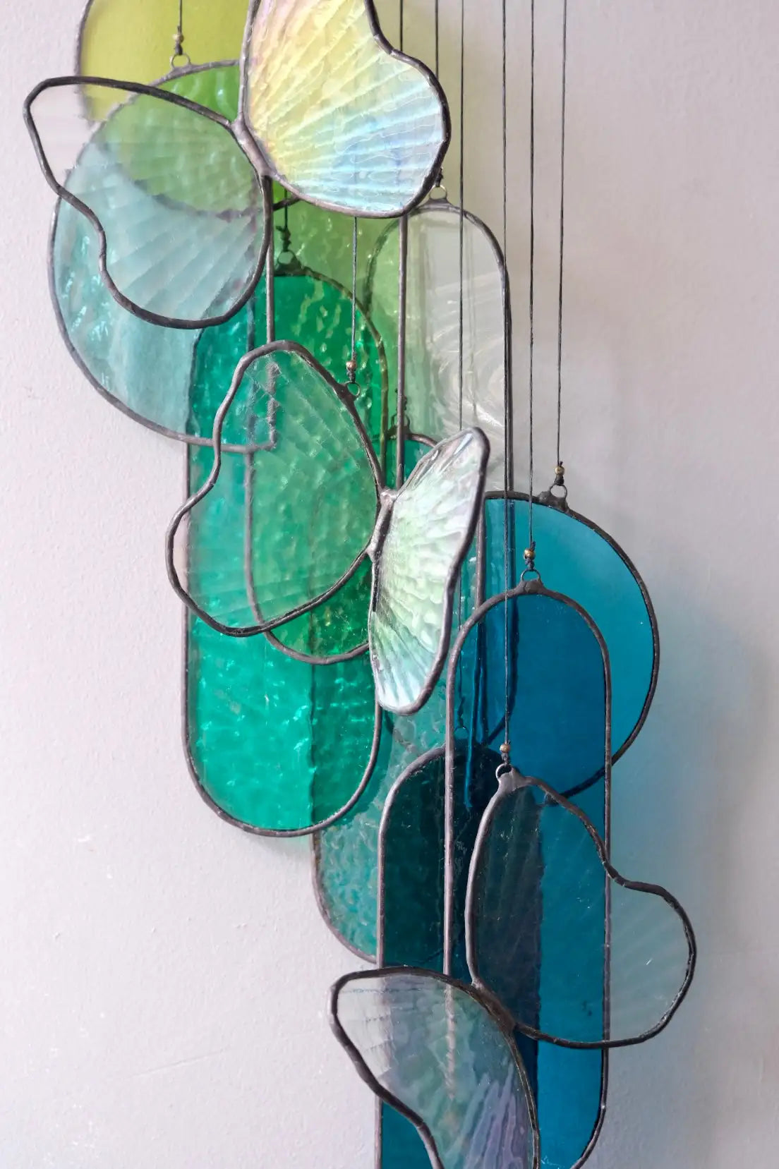 A hanging stained glass art piece featuring translucent butterfly and leaf-shaped panels in shades of green, blue, and teal, outlined with dark metal framing.