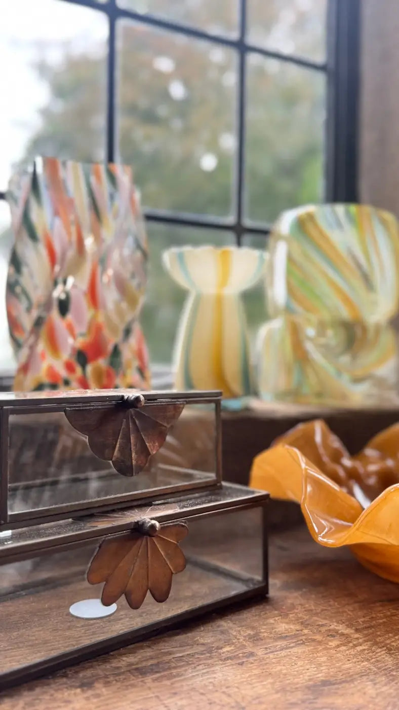 A rectangular glass jewelry box with a dark metal frame and ornate leaf-shaped handles displayed with a striped glass vase