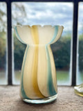 A hand-blown glass vase with a scalloped rim, featuring vertical stripes of cream, yellow, and teal blue, set against a blurred window background.