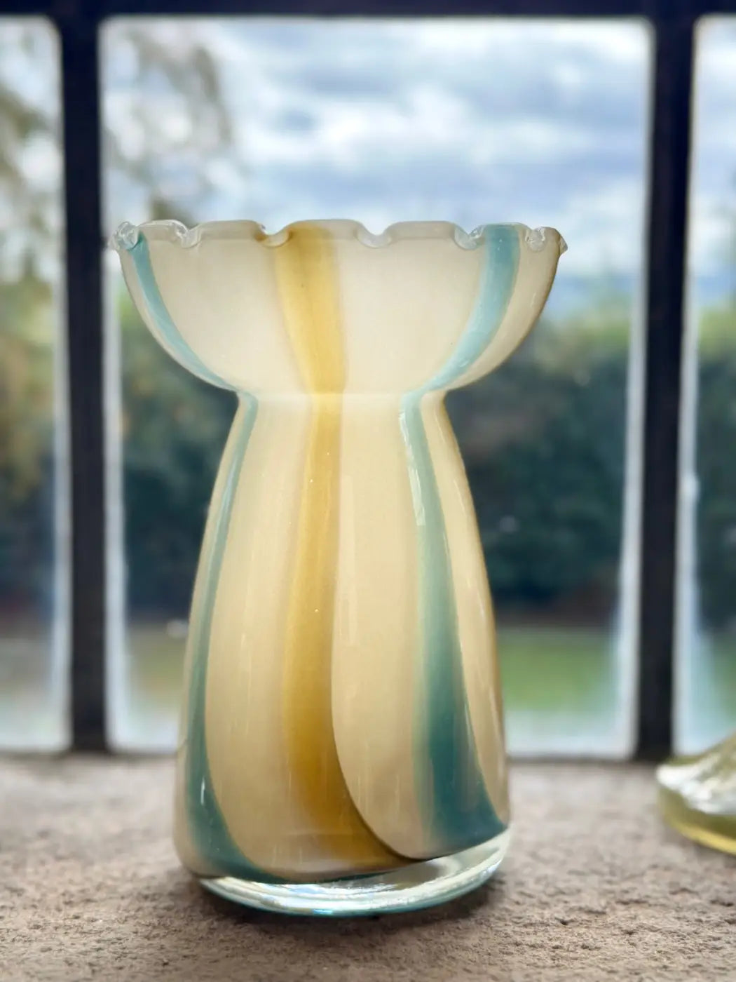 A hand-blown glass vase with a scalloped rim, featuring vertical stripes of cream, yellow, and teal blue, set against a blurred window background.