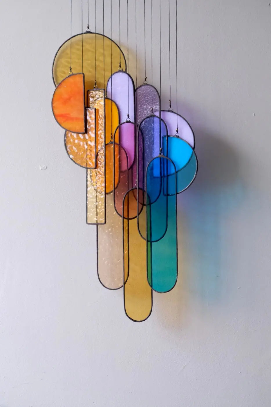 A multicolored stained glass sun catcher with various geometric shapes, hanging by strings from a brass bar, against a white wall.