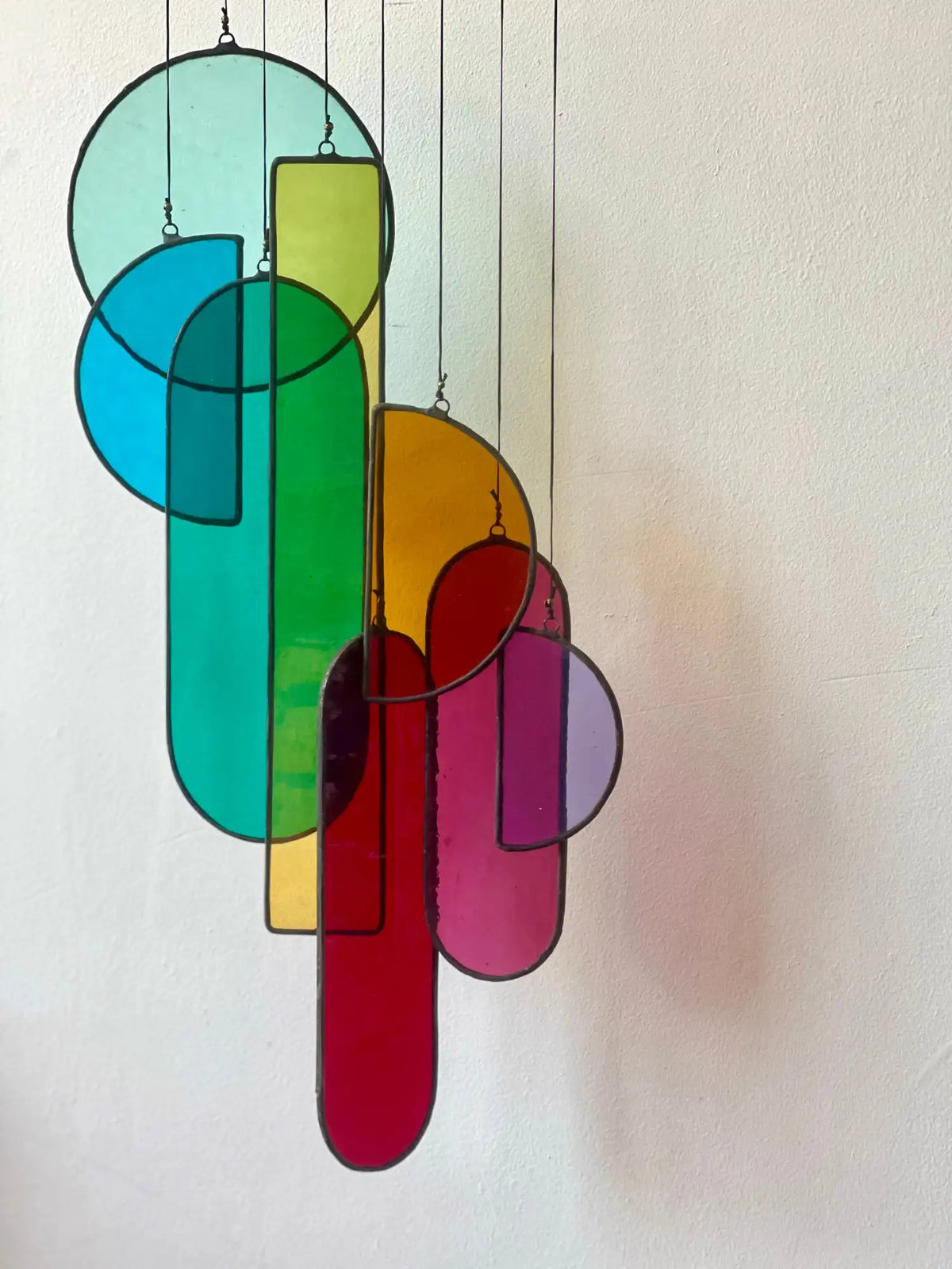 A vibrant, geometric stained glass hanging sculpture featuring overlapping circles and elongated ovals in translucent hues of teal, green, yellow, orange, red, and purple.
