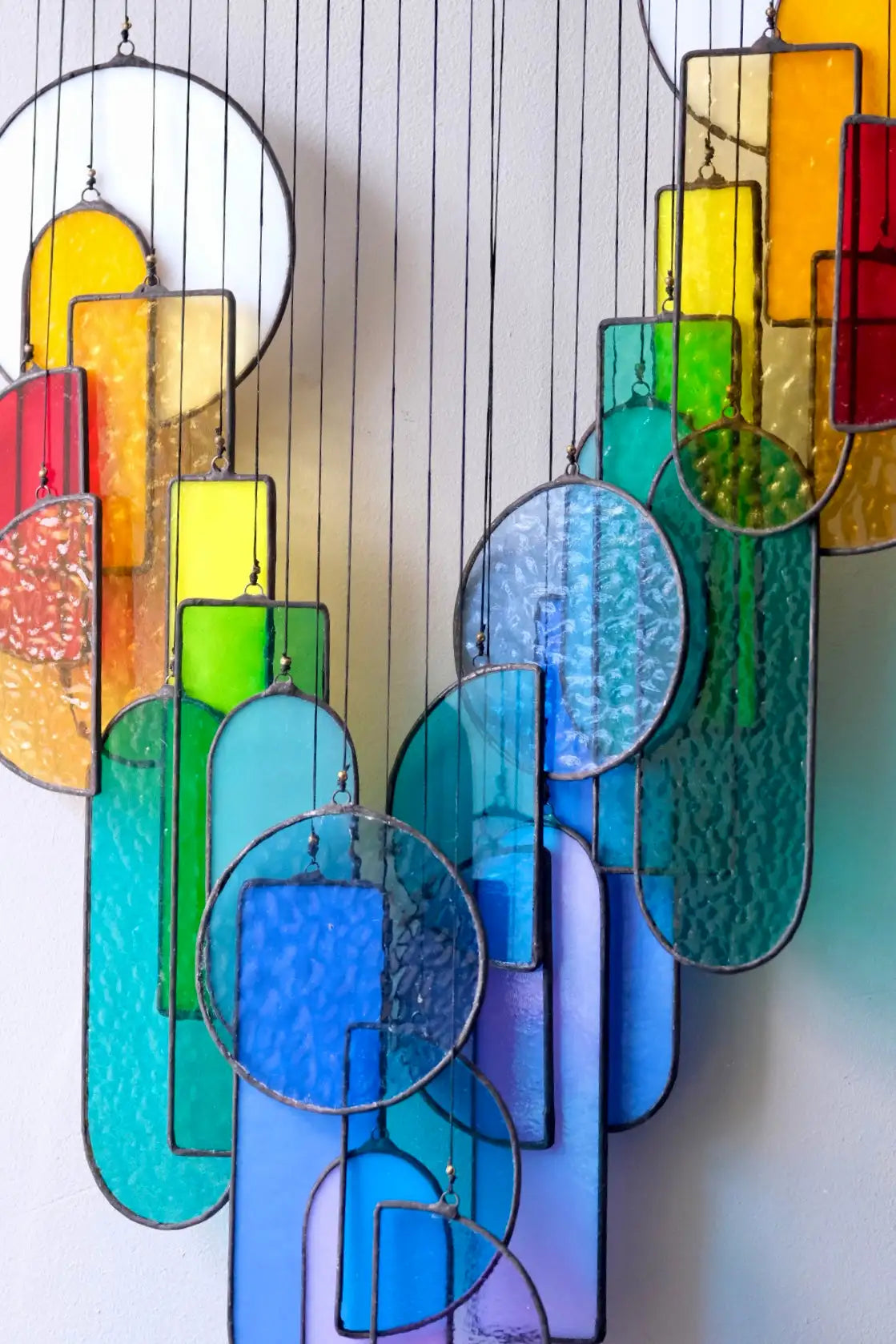 A vibrant, rainbow-colored stained glass hanging art piece featuring geometric shapes and textured panels suspended by thin wires.