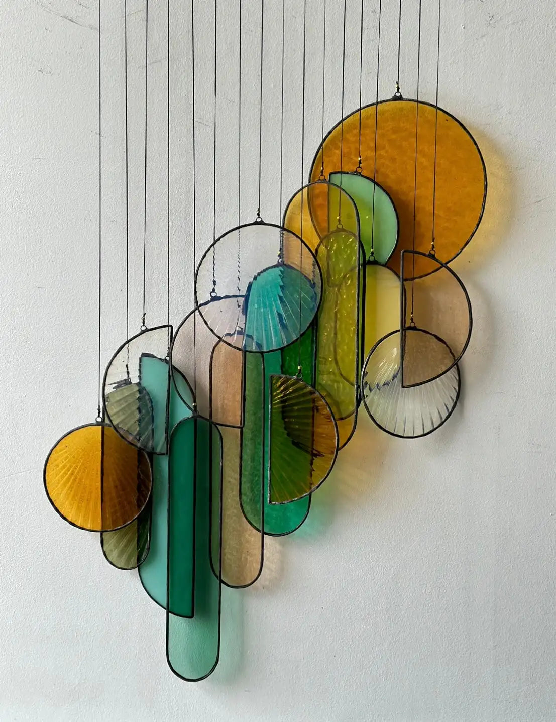 A vibrant stained glass hanging mobile featuring circular and elongated shapes in shades of amber, teal, green, and yellow, outlined with dark metal frames.